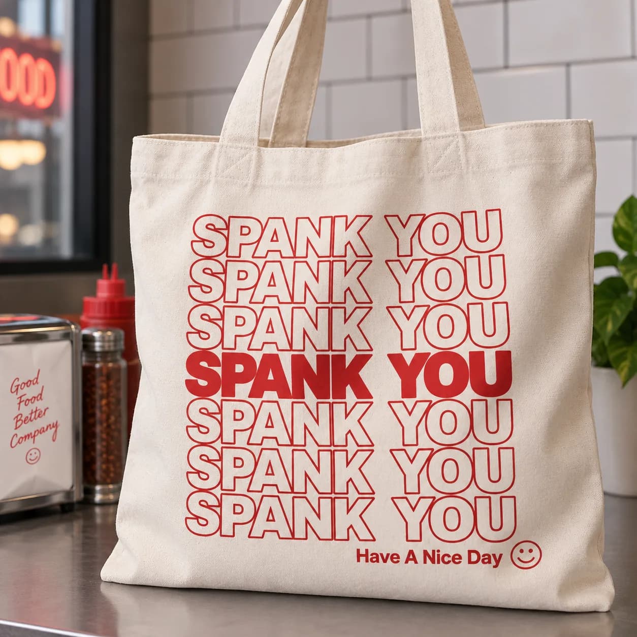 A natural canvas tote bag on a diner counter, printed with SPANK YOU repeated seven times in red — the middle line solid, the rest outlined — and Have A Nice Day with a smiley in the corner.